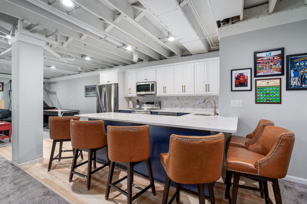 Basement renovation kitchen addition with two-tone colors in dark navy cabinets below and bright white cabinets above. A white granite countertop and six brown bar stool chairs sit around the bar.