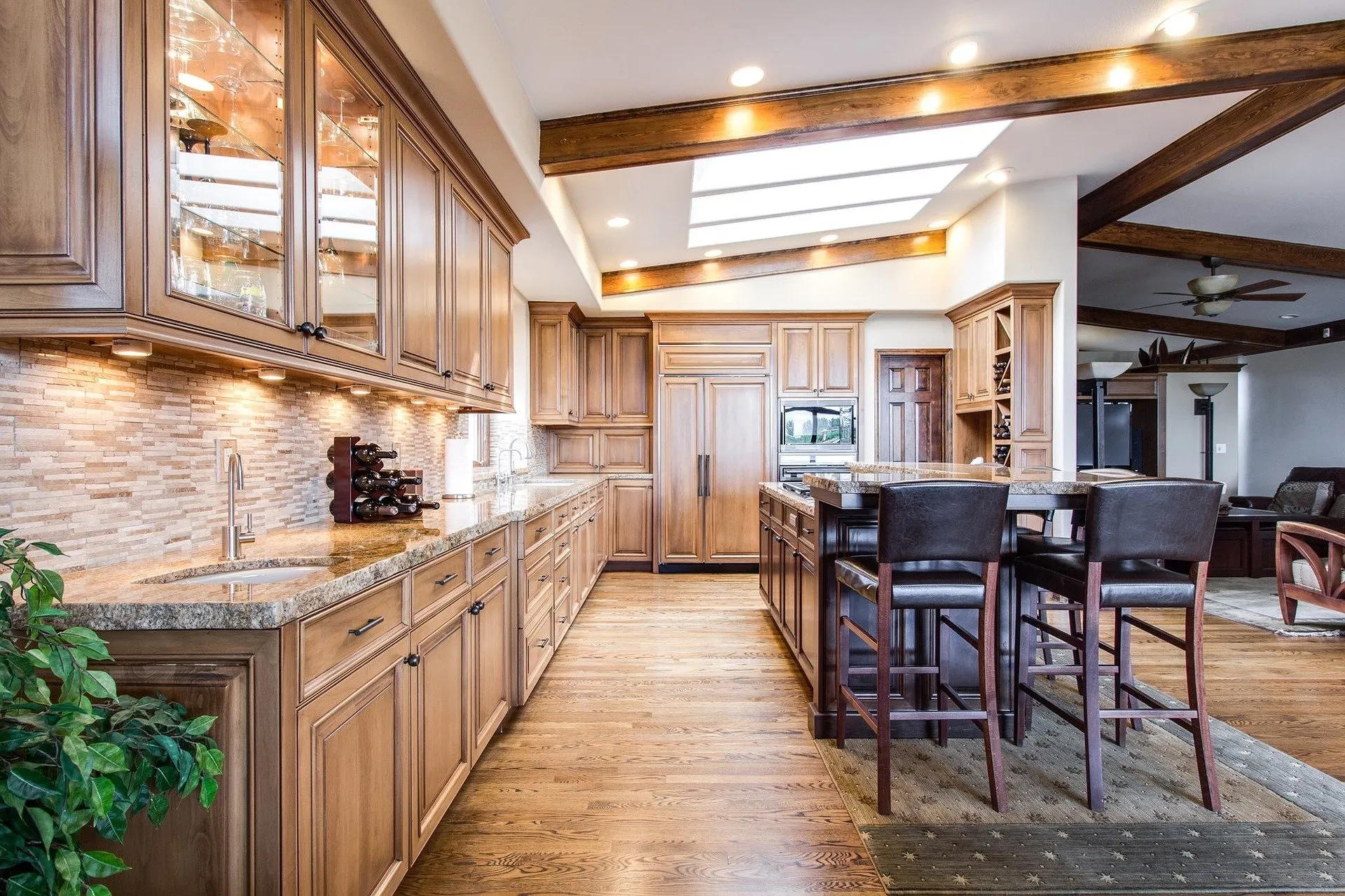 Kitchen with wood cabinetry, including a long counter topped with marble and display hutch with glass windows, kitchen island with breakfast bar chairs and open floor plan leading into living room space