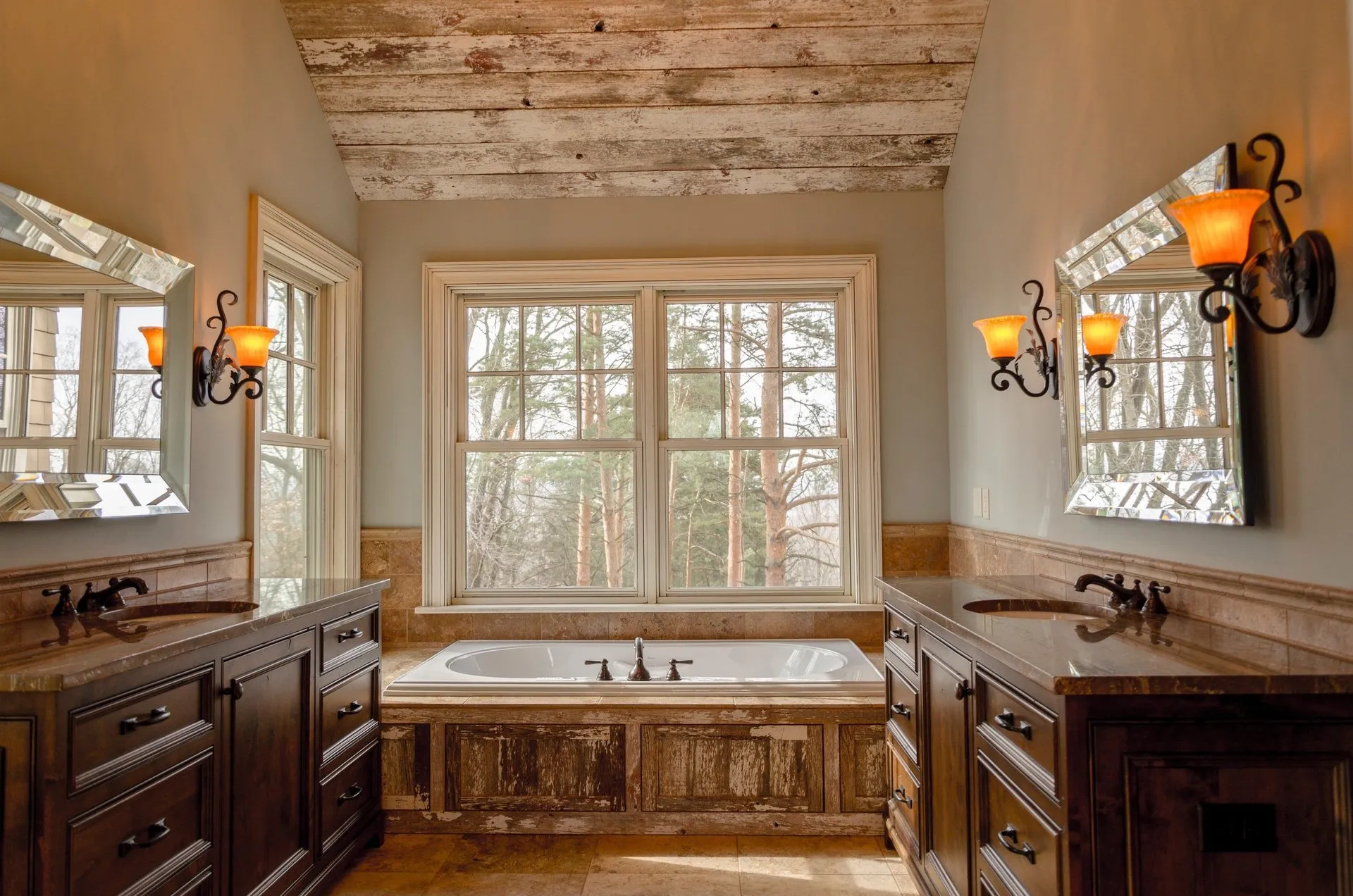 Rustic bathroom with full tub, two bathroom sink vanities face each other with two matching mirrors above each one. Decorative lighting fixtures hang on either side of both mirrors.